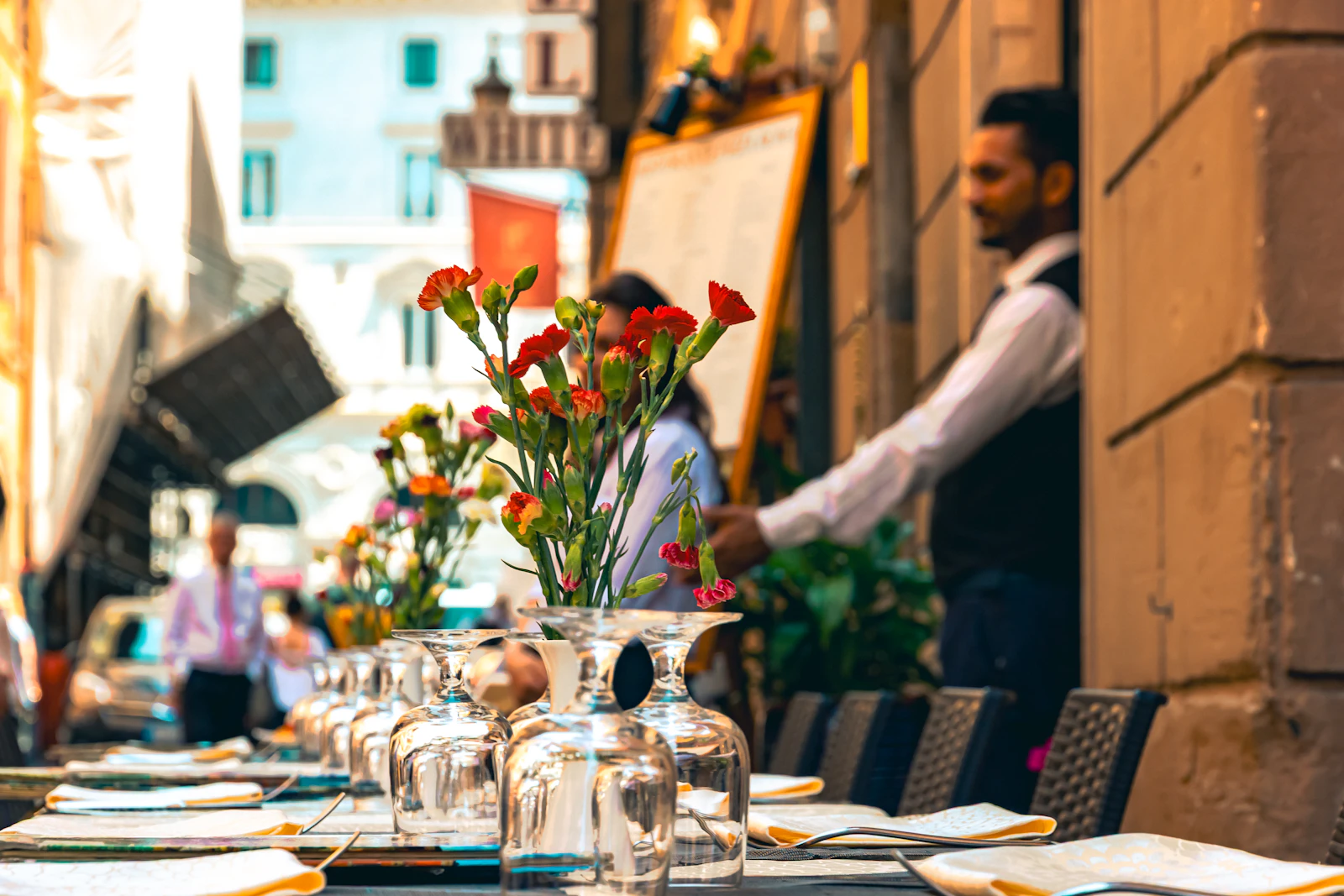 Outdoor restaurant table set with glassware and menus while a server moves through service